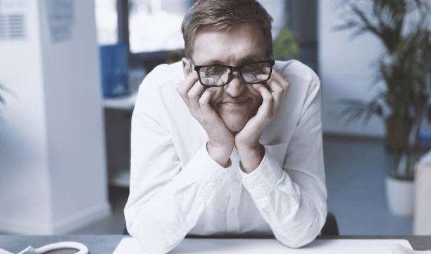 The image features a smiling man resting his chin on his hands. He is wearing square-framed glasses and a white shirt. He is sitting at a desk in a well-lit office environment with a relaxed and cheerful demeanor.