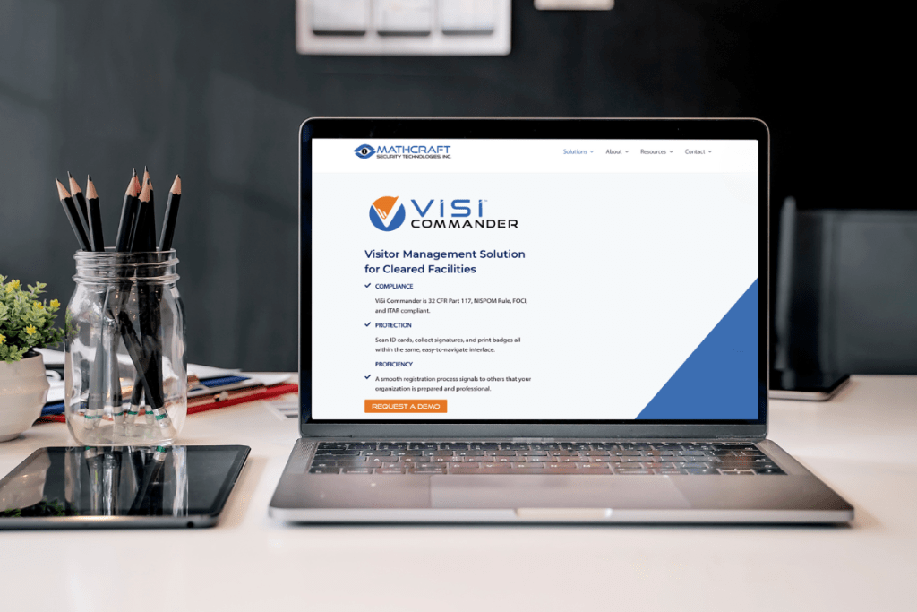 A laptop is open on a desk displaying the homepage of MathCraft Security Technologies Inc. The screen shows information about 'ViSi Commander,' a visitor management solution for cleared facilities, highlighting its compliance with security regulations, protection features, and proficiency. A section labeled 'REQUEST A DEMO' is visible. The laptop is on a tidy desk with a glass jar filled with pencils, a small potted plant, and a tablet reflecting the laptop screen, suggesting a professional and organized workspace