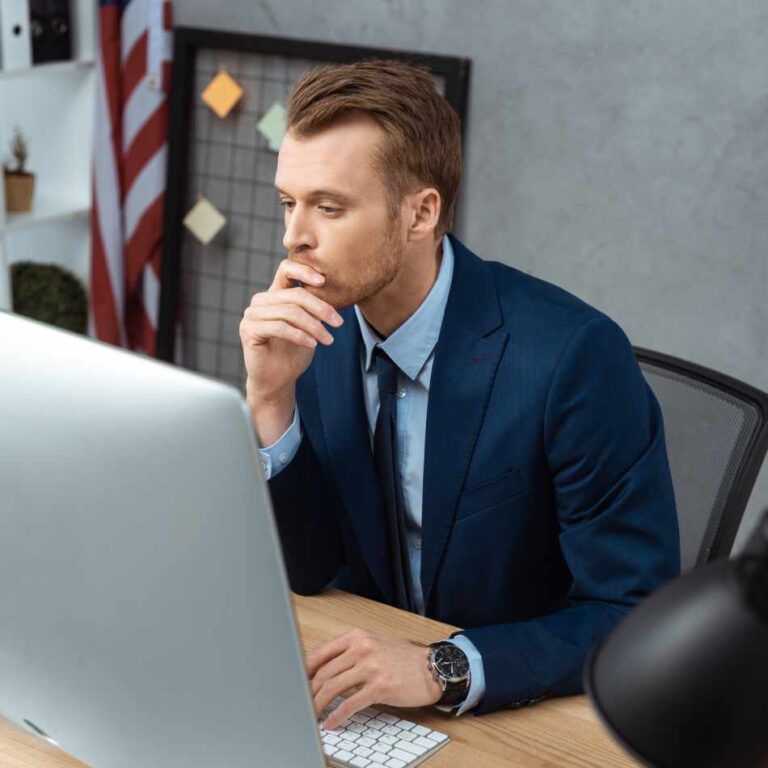 Photo of FSO consultant looking at computer managing multiple clients, the United States flag in the background. His hand is on the keyboard and his hand is in his mouth thinking.
