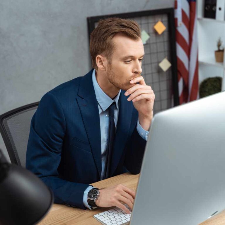 Photo of FSO consultant looking at computer managing multiple clients, the United States flag in the background. His hand is on the keyboard and his hand is in his mouth thinking.